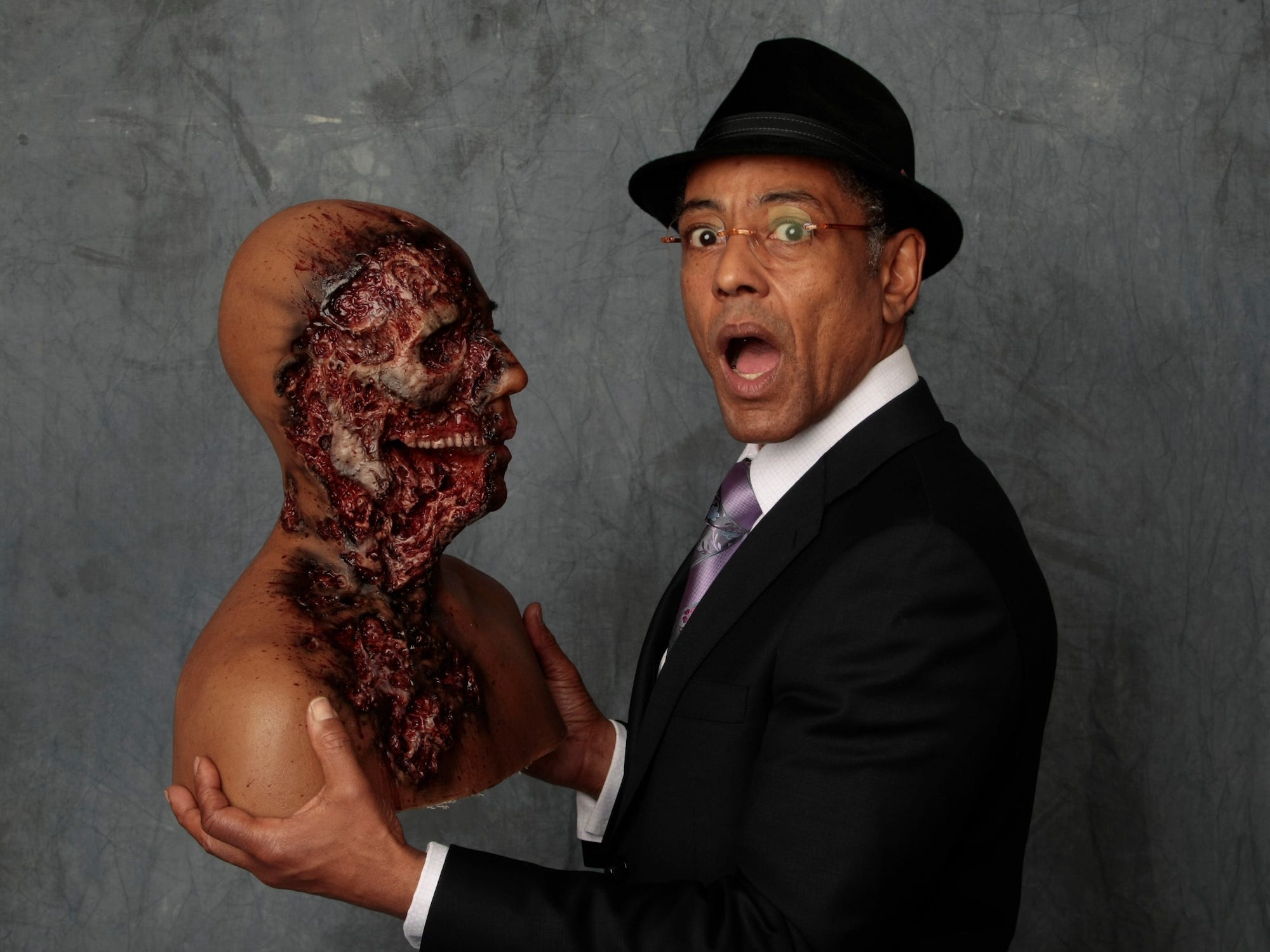 Actor Giancarlo Esposito pretends to look shocked while holding a bust of his head with a large chunk of its face missing