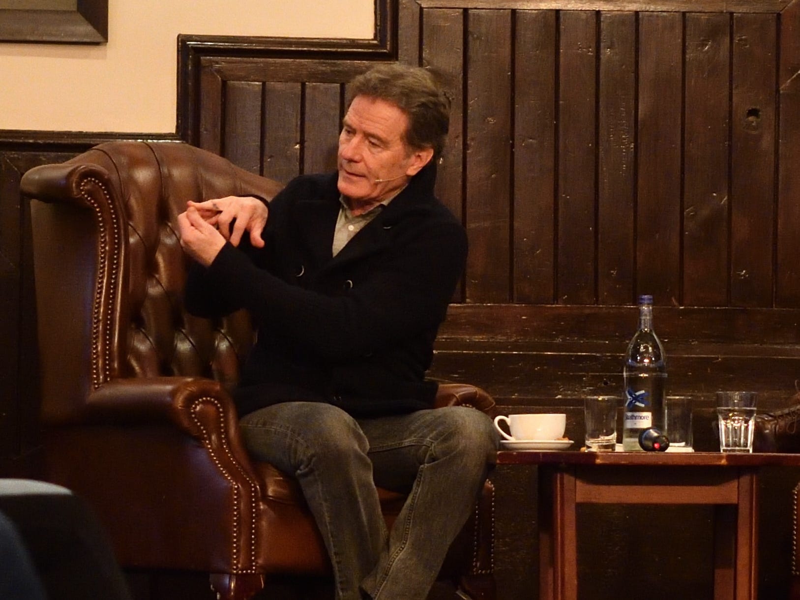 Bryan Cranston shows his finger which has a Breaking Bad chemical element tattoo while sitting in a brown leather chair