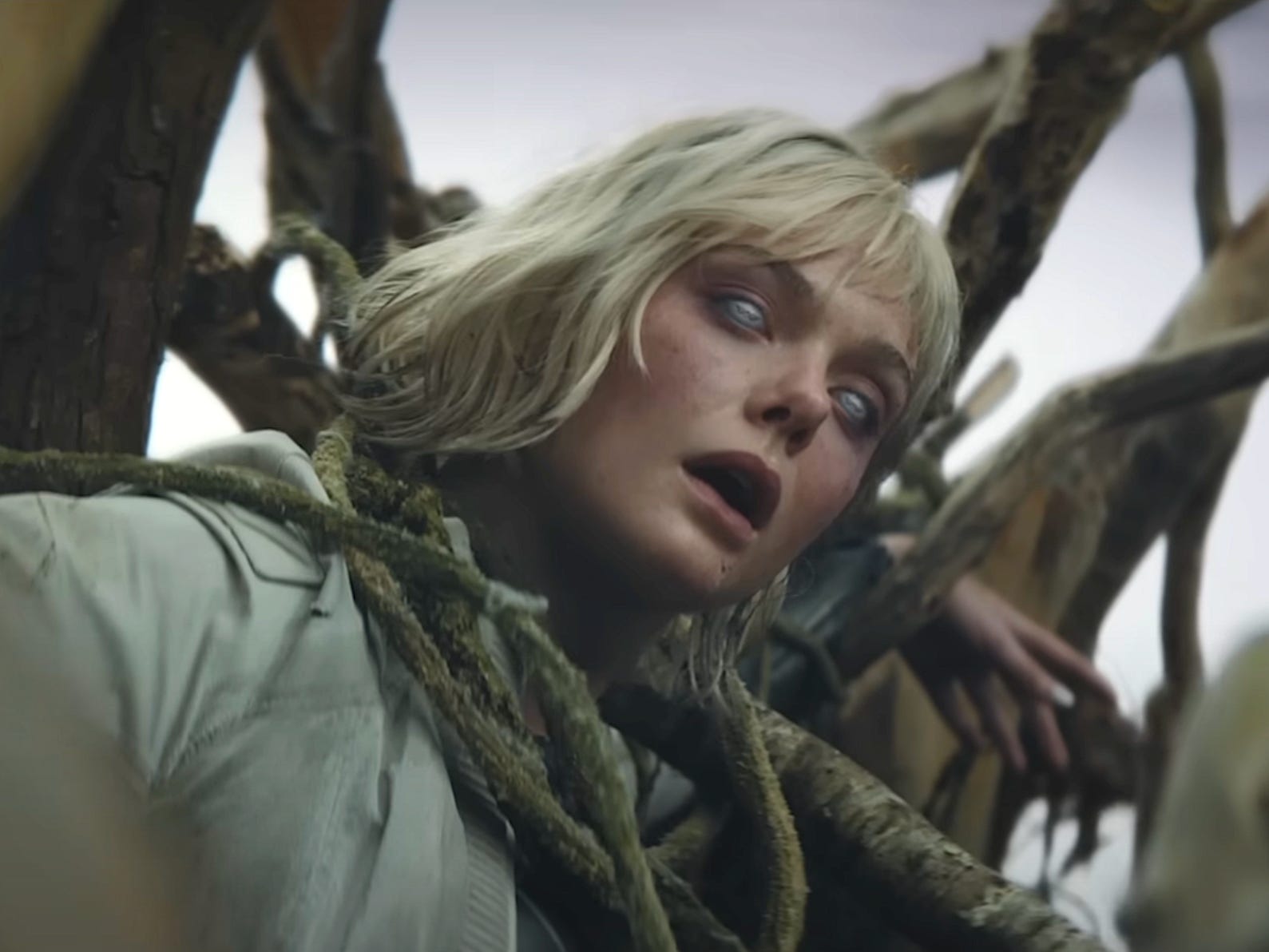 A young woman with short blond hair is restrained by branches in a tree. She has her mouth open and her eyes are silver with a black 'W' logo on them.