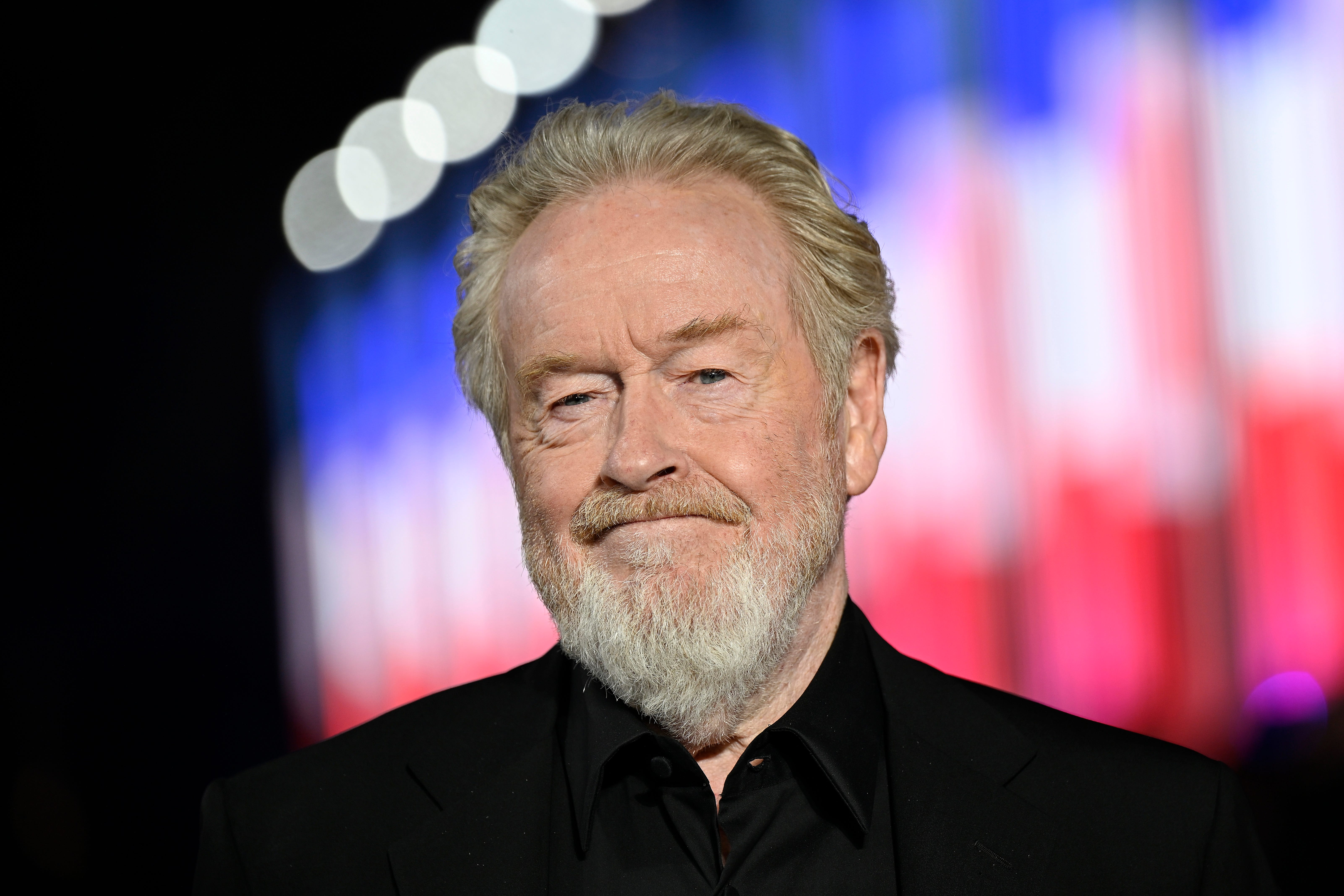 Ridley Scott is shown smilling in front of a red white and blue blurred background.