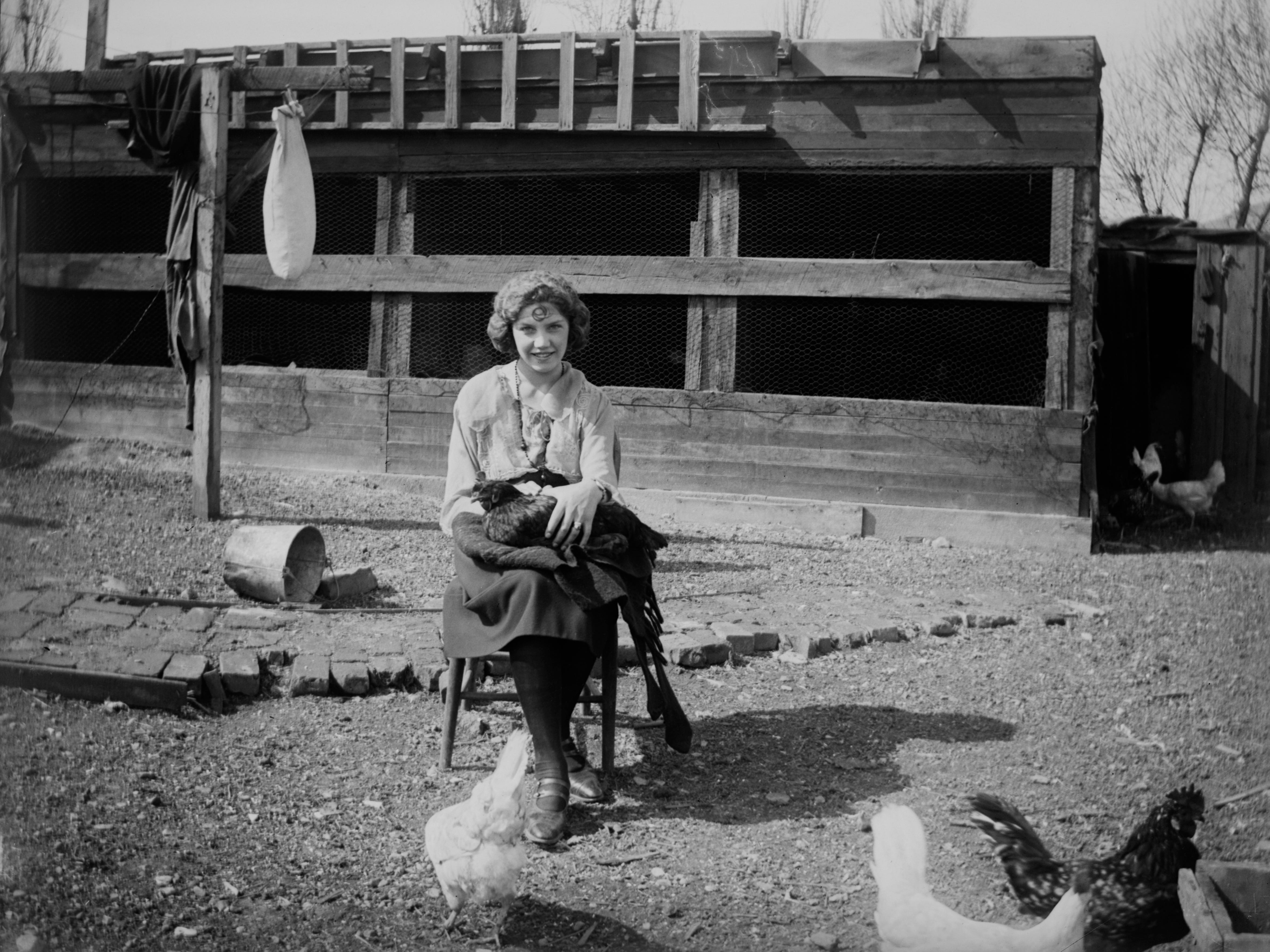 A woman holding a chicken while sitting on a stool in front of a chicken coop, 1925. Other chickens are seen on the ground.