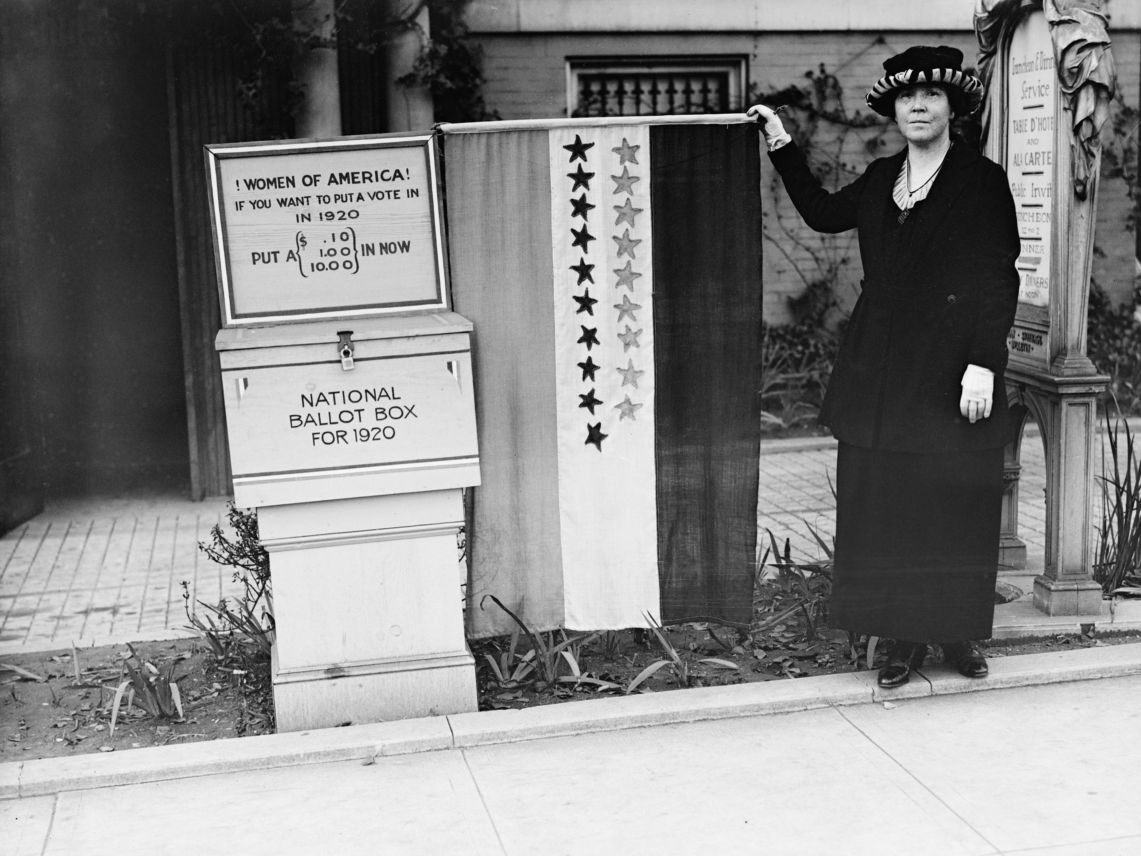 A suffragette standing next to a National Ballot Box in 1920. Above the ballot box is a sign that reads, "Women of America! If you want to put a vote in in 1920 put a ($0.10, 1.00, 10.00) in now." She also appears to be holding a flag.