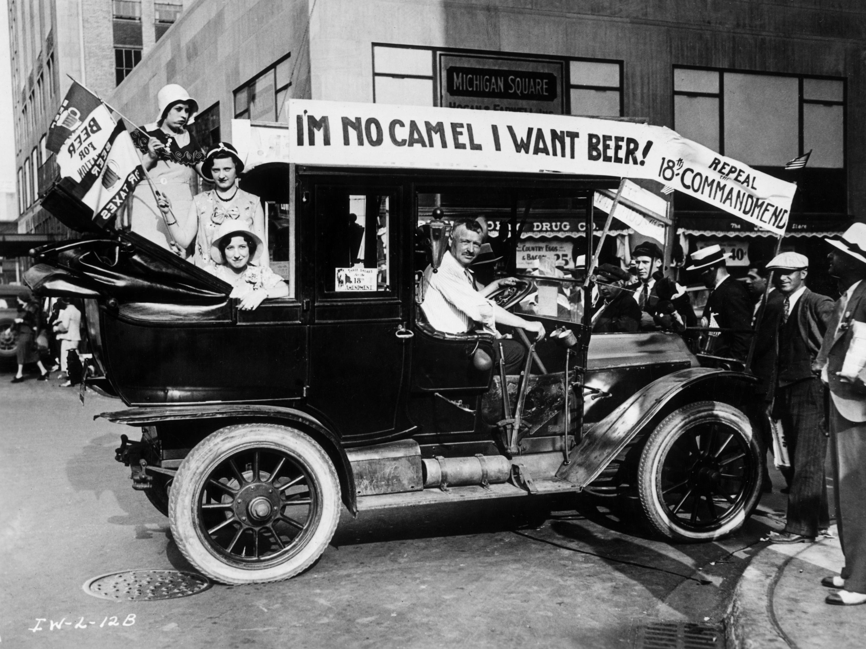 A group of people in a car protesting Prohibition. The car is decorated with a sign that reads, "I'm no camel I want beer!"