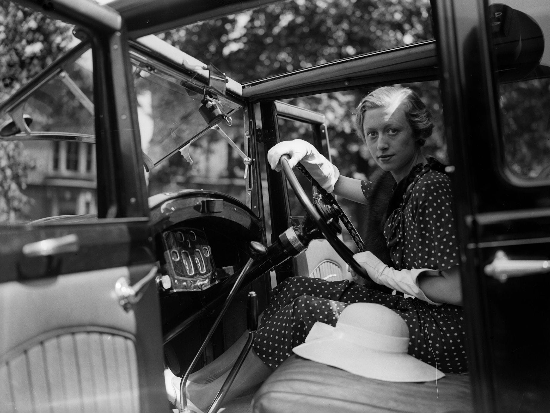 A woman sitting in a car wearing a polka dot dress. A wide-brimmed hat sits on the seat next to her.