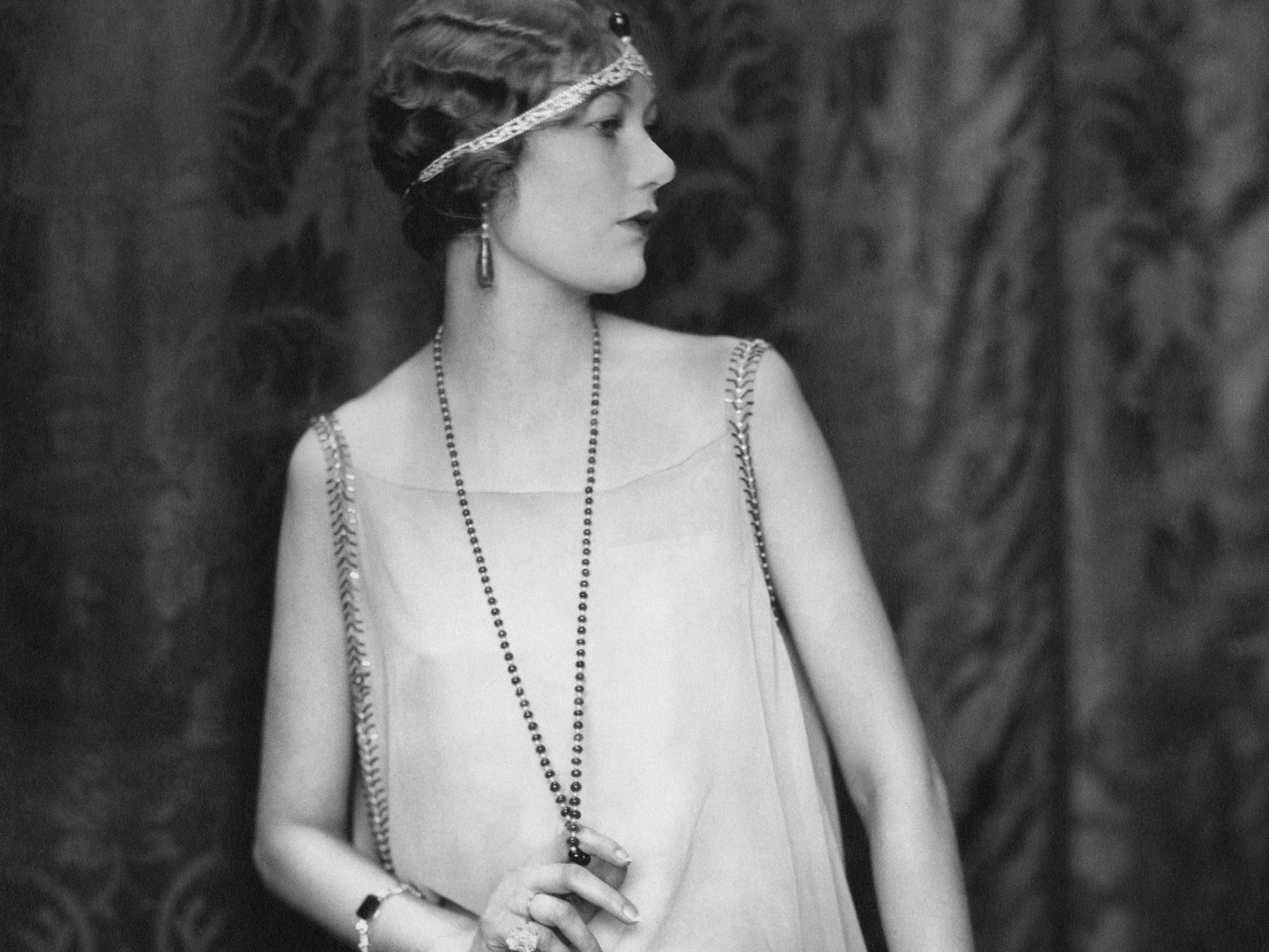 A woman in the 1920s shows off the decade's classic fashion, which included a headband, loose dress, and long chain necklace..