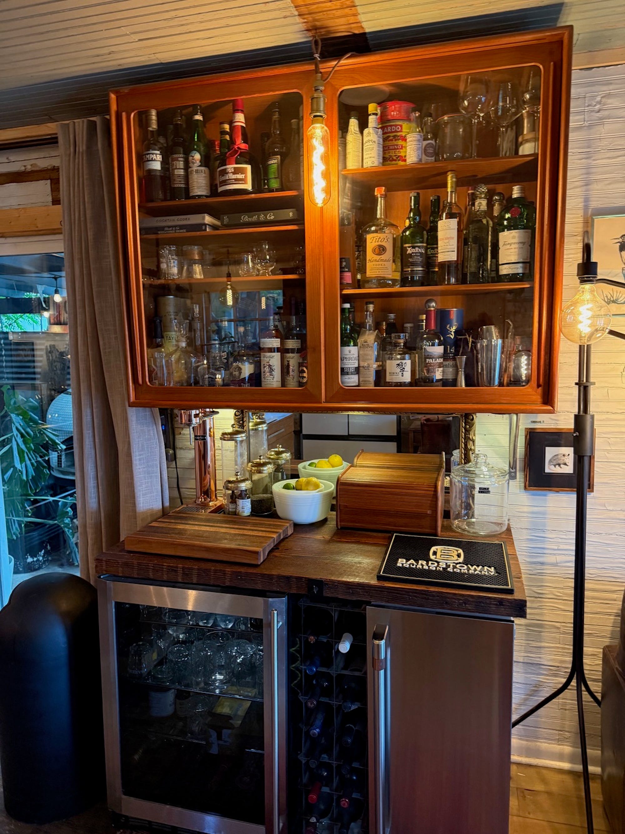 A bar with a mini fridge and shelving above it with a glass cover.