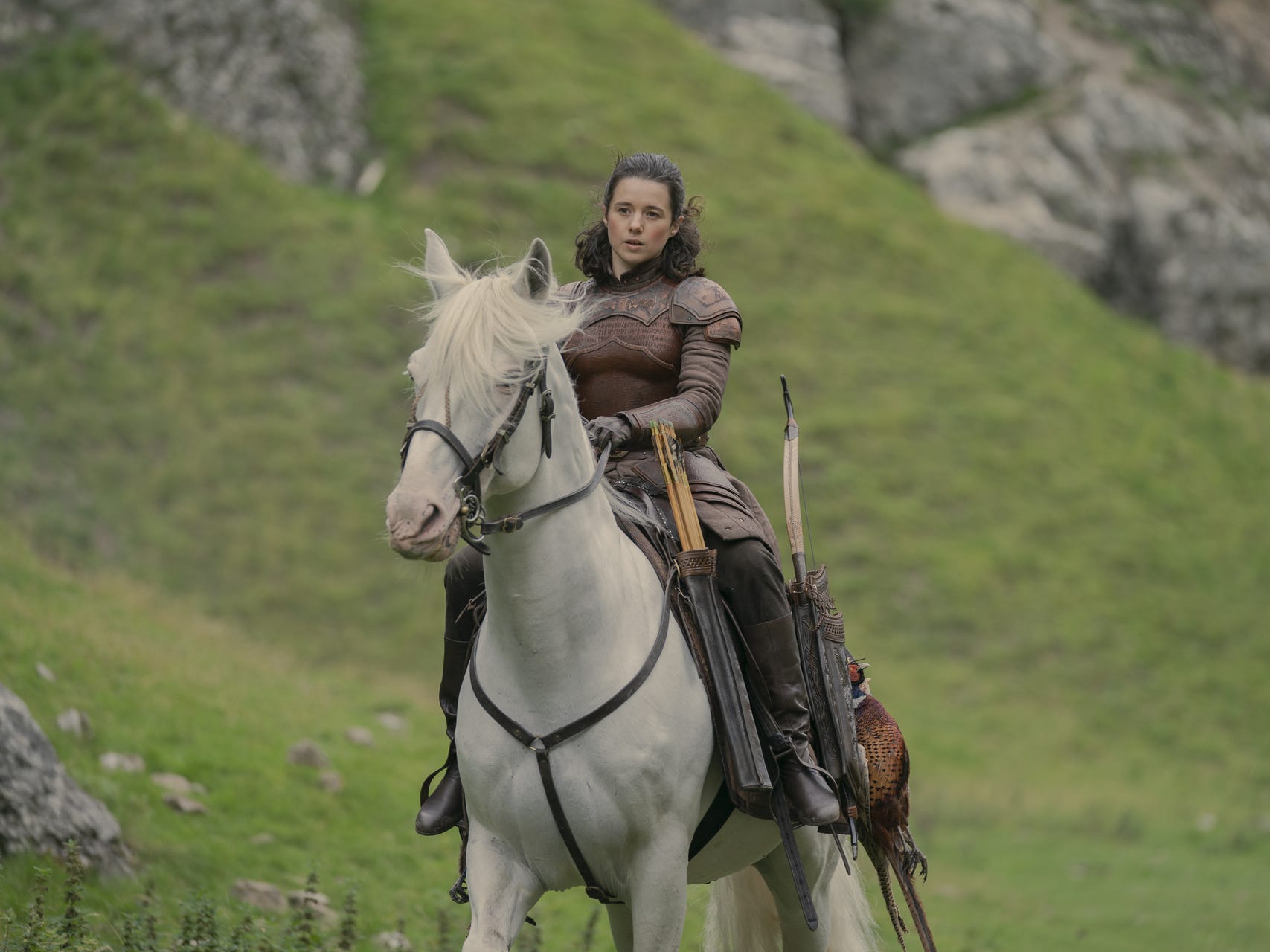 rachel redford as lady rhea royce in house of the dragon, a young woman with brown hair pulled half back, wearing brown armor and sitting astride a white horse, riding through a grassland