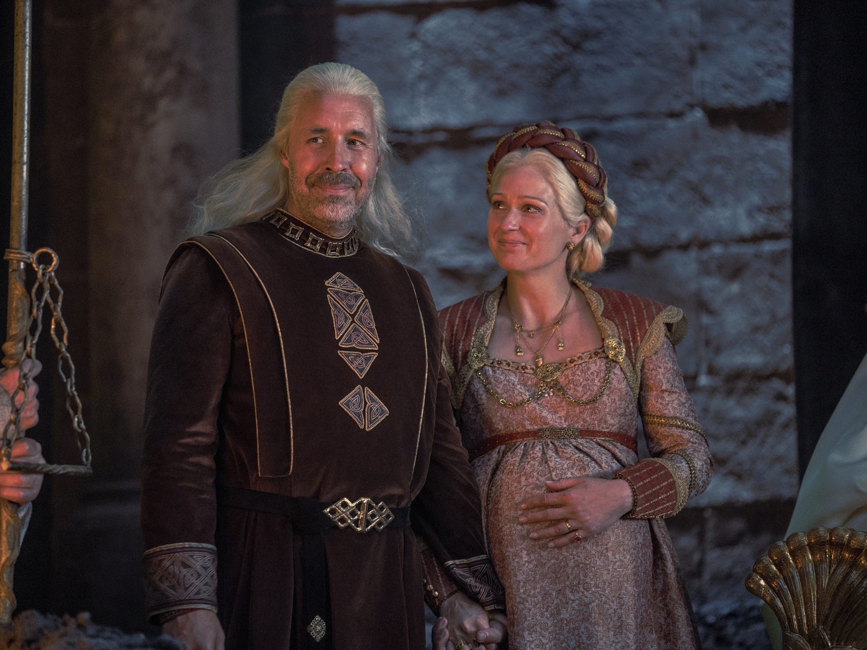 A man and woman with silver hair wearing medieval royalty garb stand lovingly next to each other.