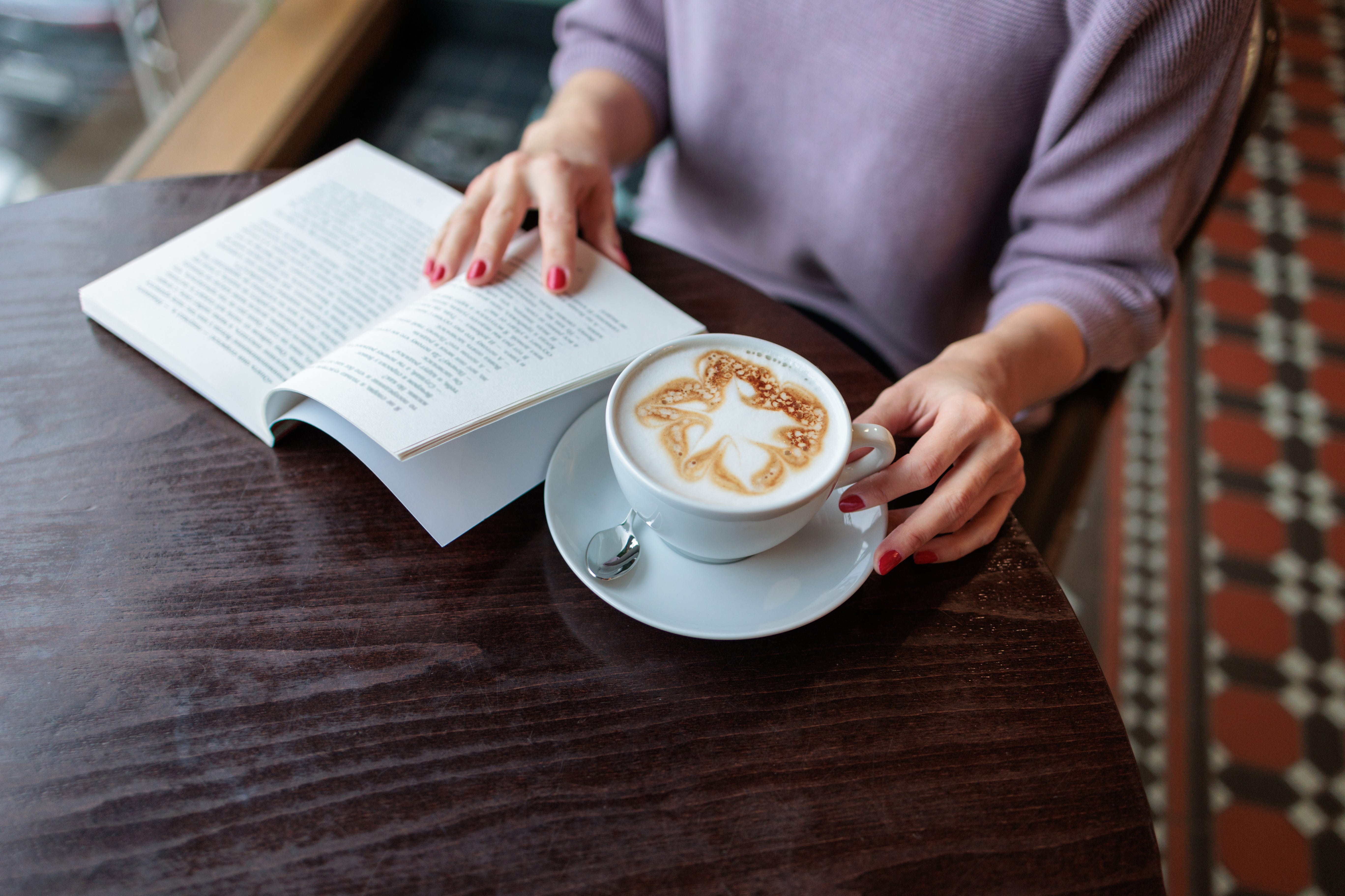 Woman holding book open while grabbing cappucino on table