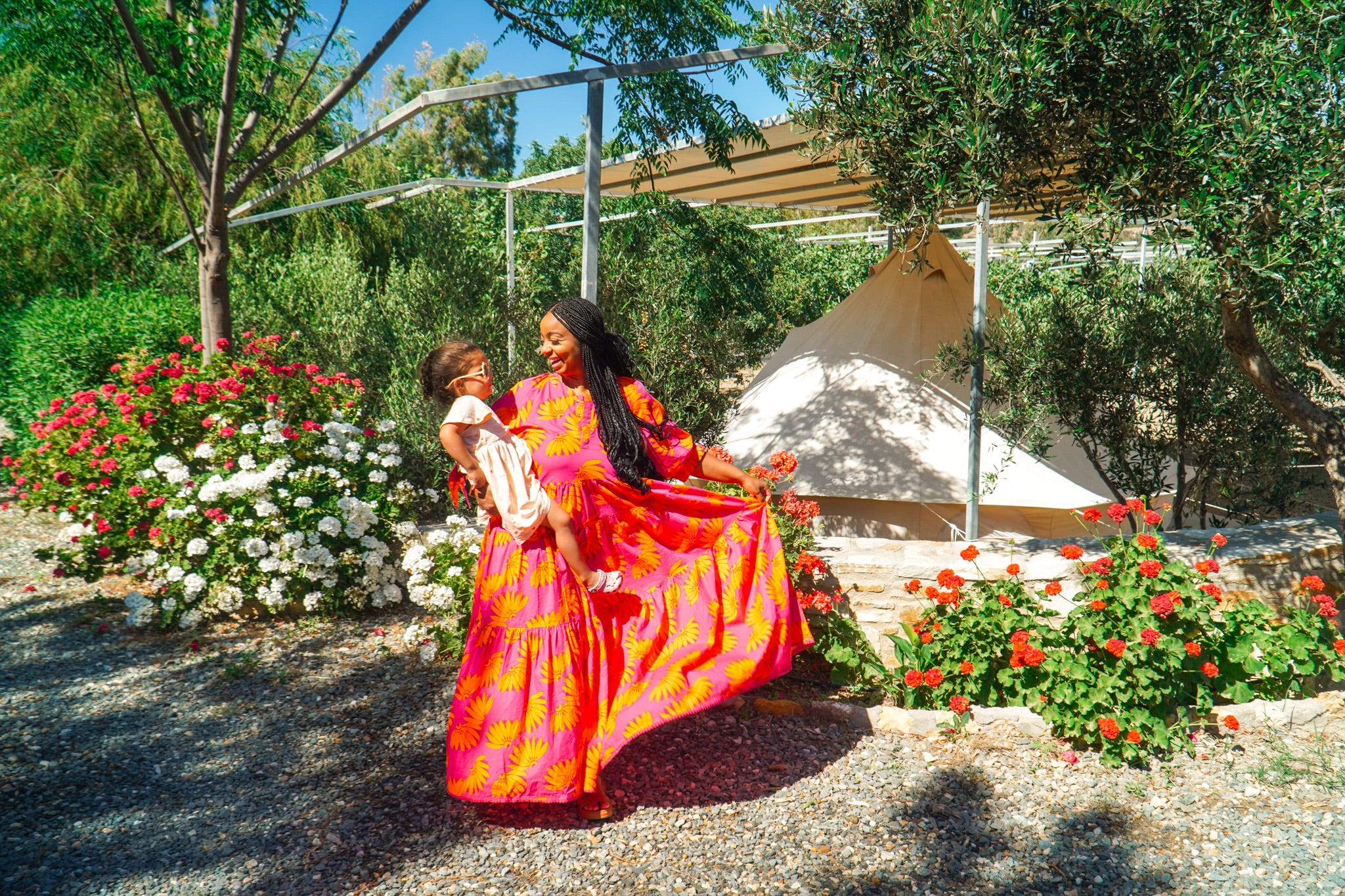 Adult in bright floral dress holds a child in a sunny garden with flowers and a canvas tent.