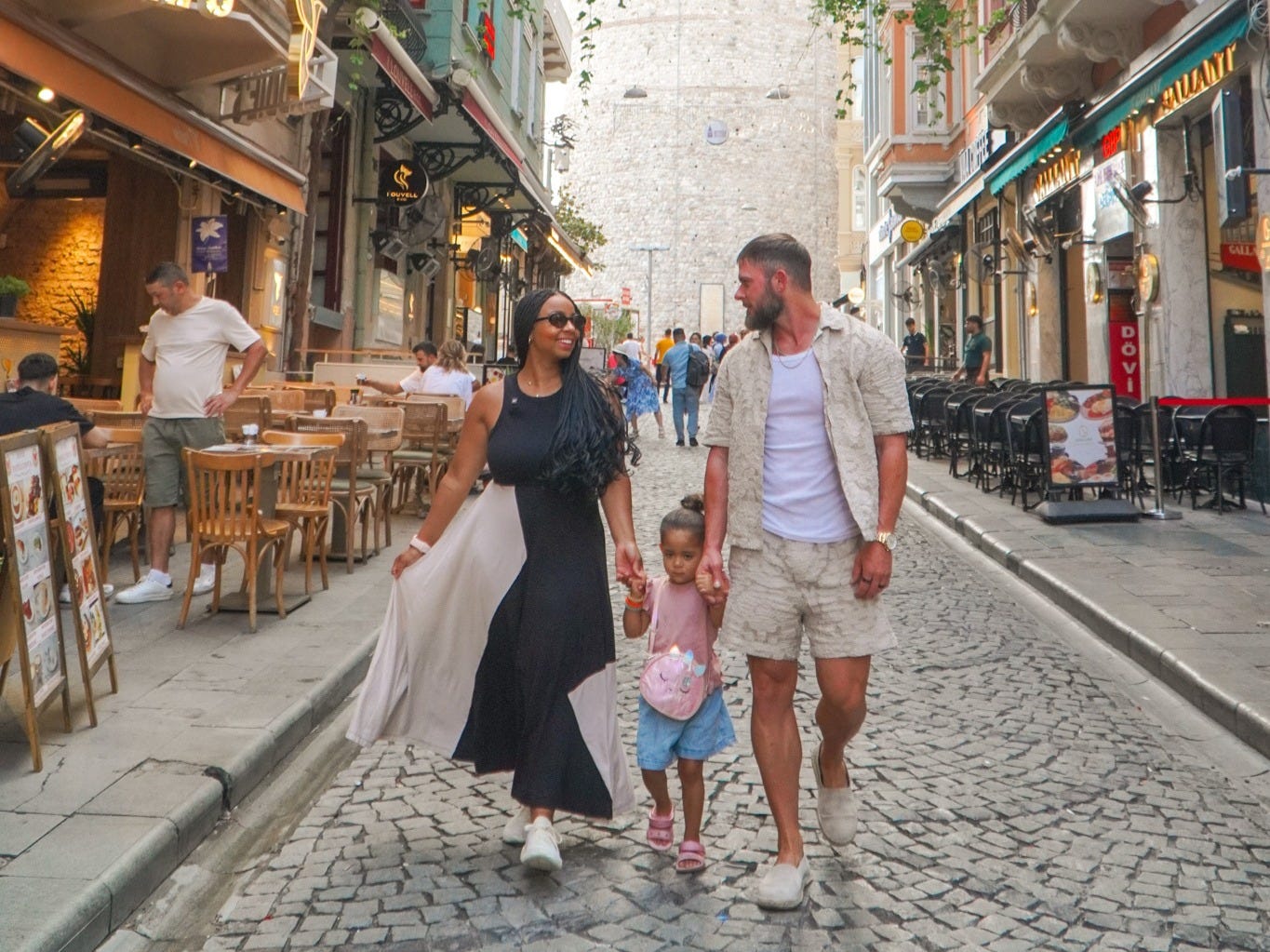 Three pedestrians walk along a cobblestone street with cafes beneath the Galata Tower in Istanbul.