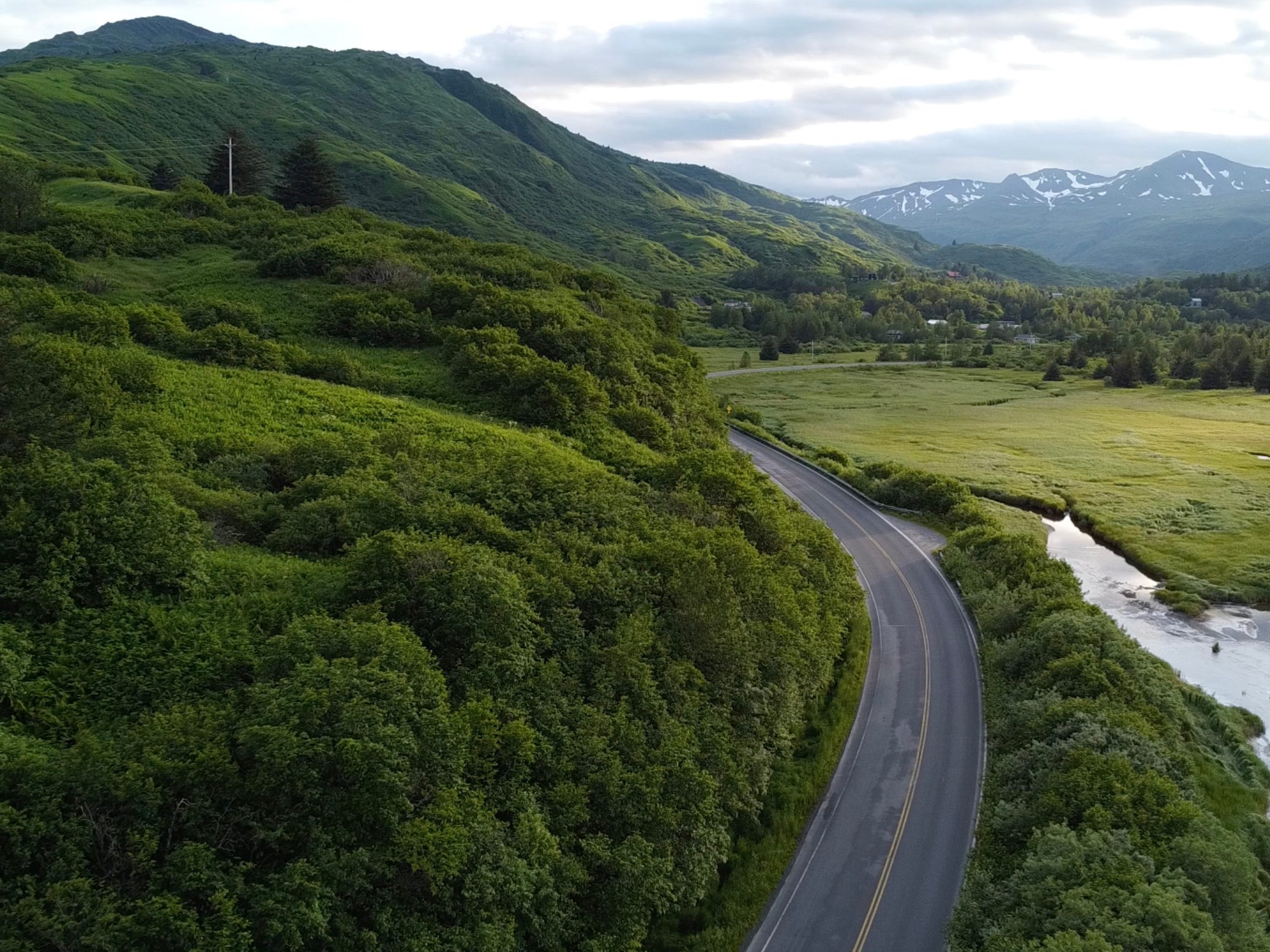 A winding road runs through a green mountain valley beside a narrow river in remote Alaska.