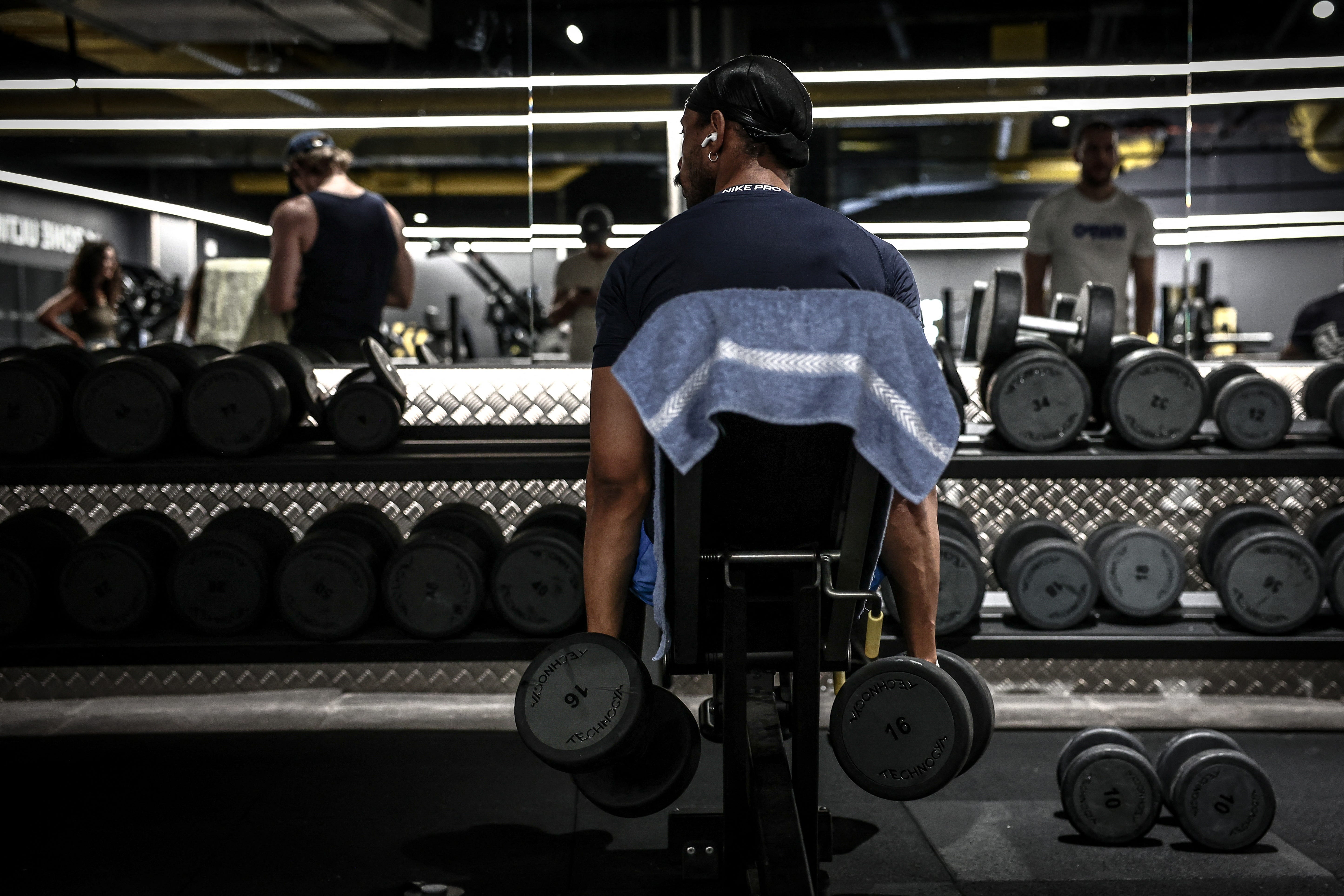 Person seated at a gym bench holds dumbbells in front of a mirrored weight rack.