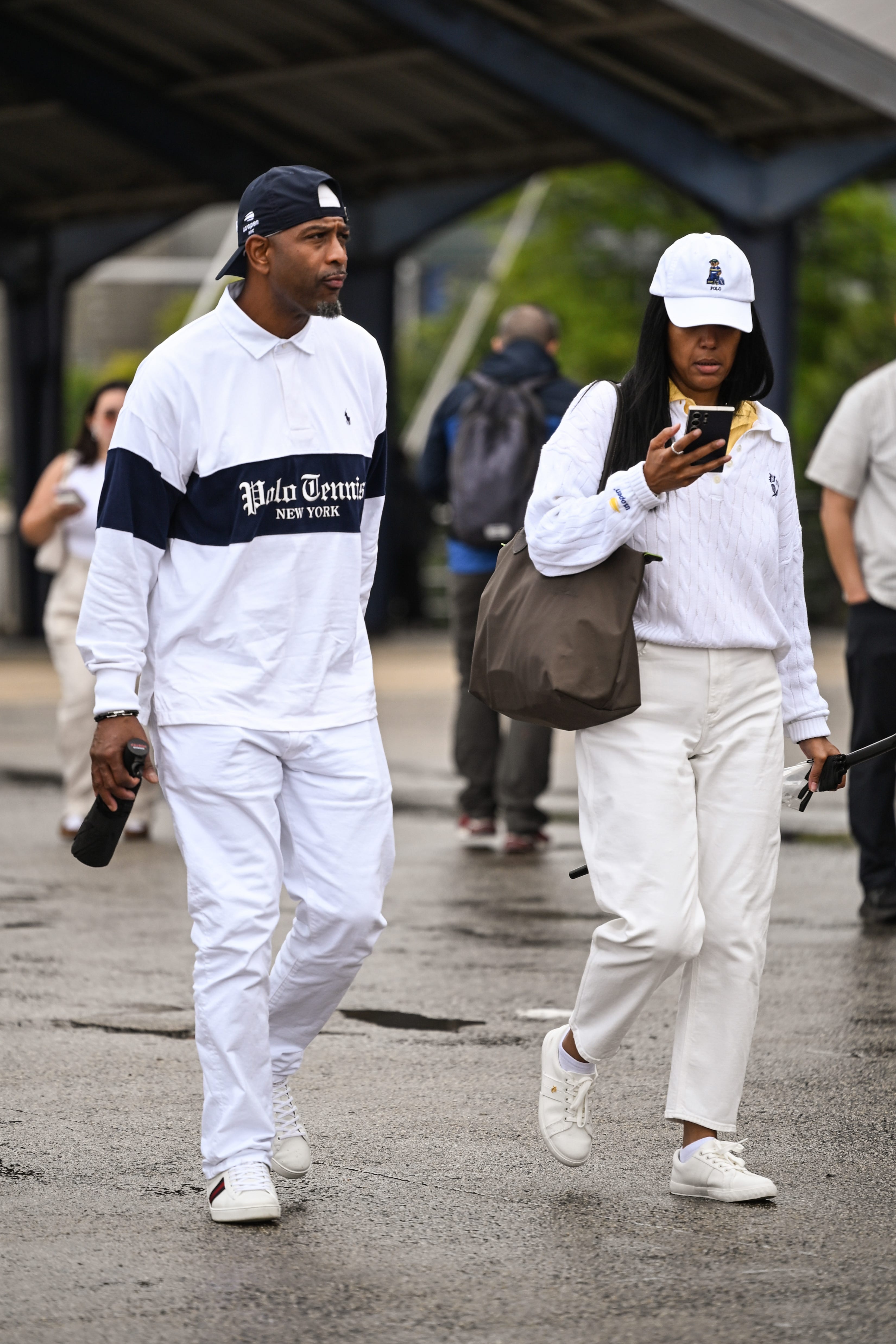 Two people in coordinated white Polo Tennis outfits walk on wet pavement, one looking at a smartphone.