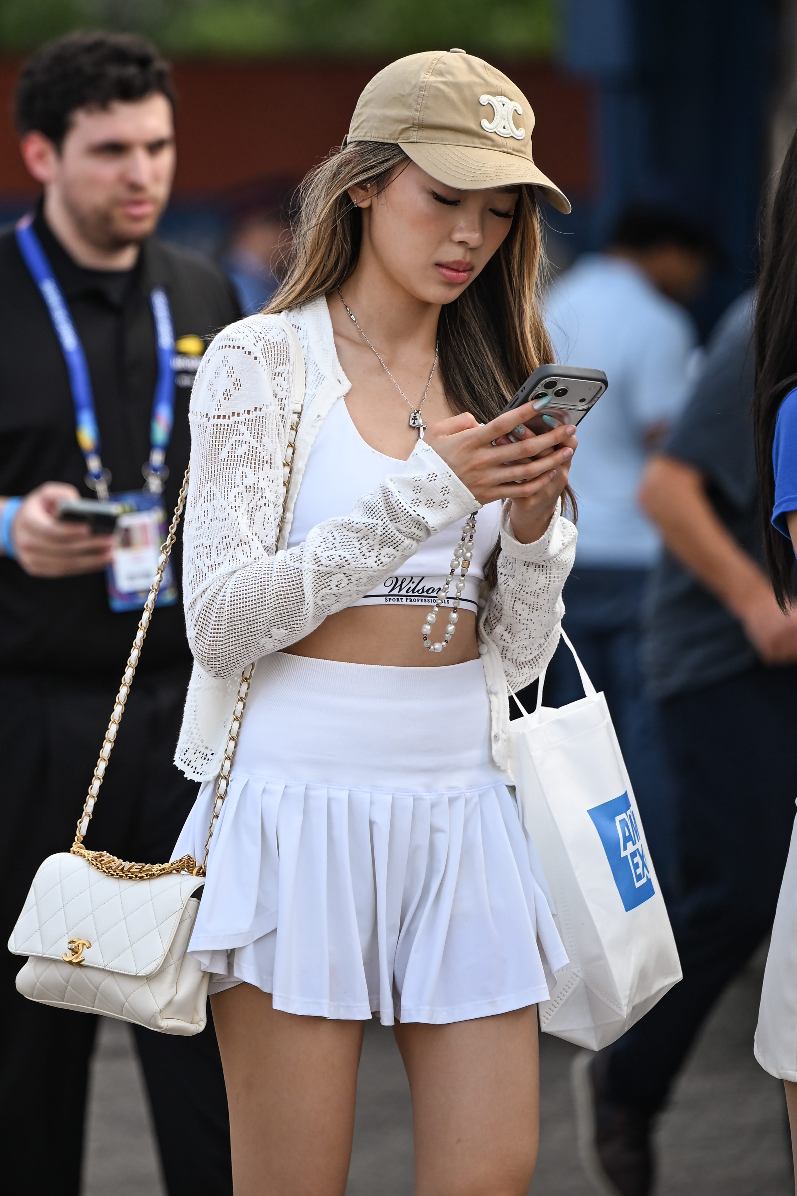 Person in a white tennis-style outfit and beige logo cap looks at a smartphone while carrying a quilted handbag.