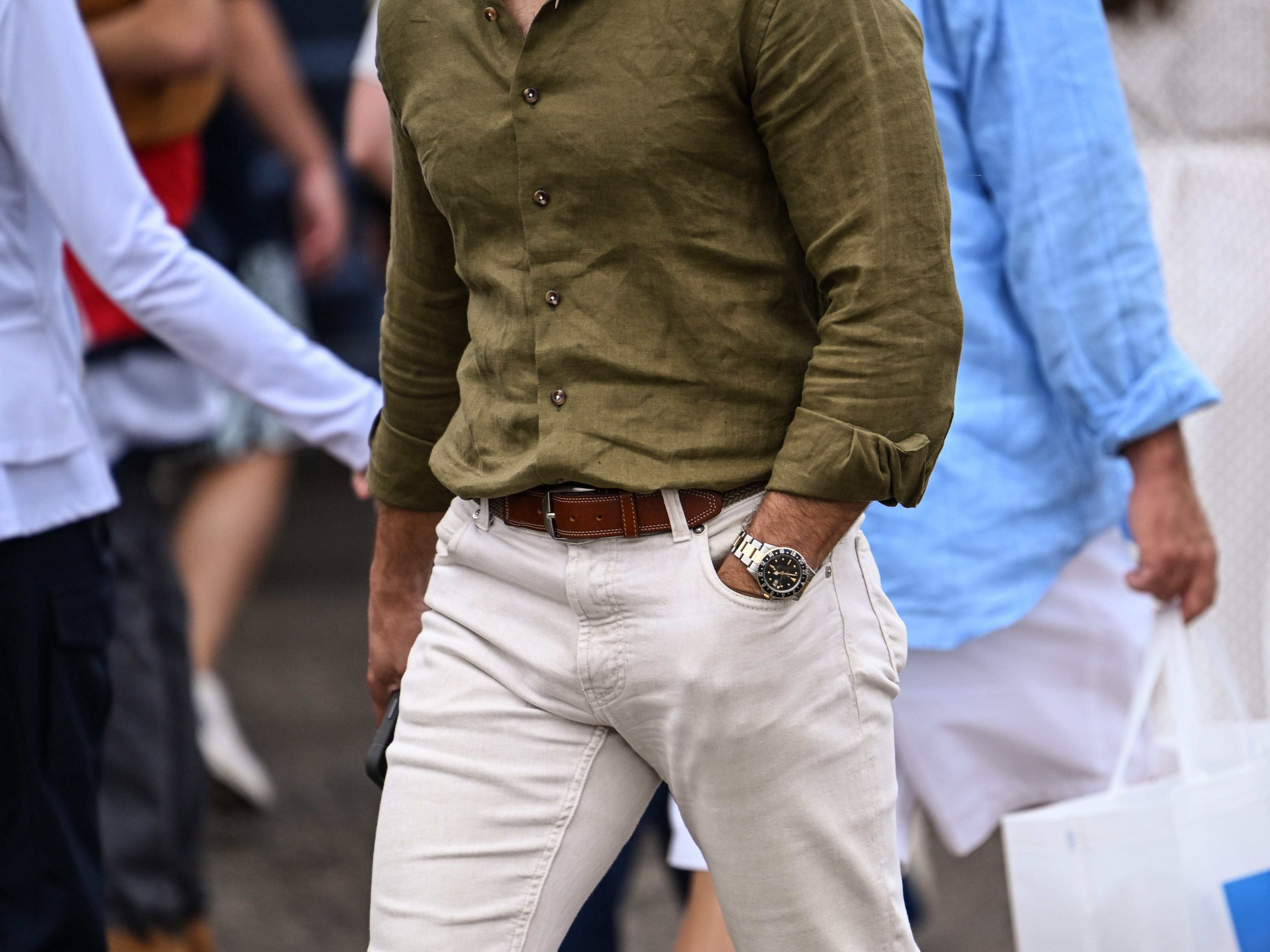 Person in an olive shirt and light trousers walks through a crowded street wearing a wristwatch.