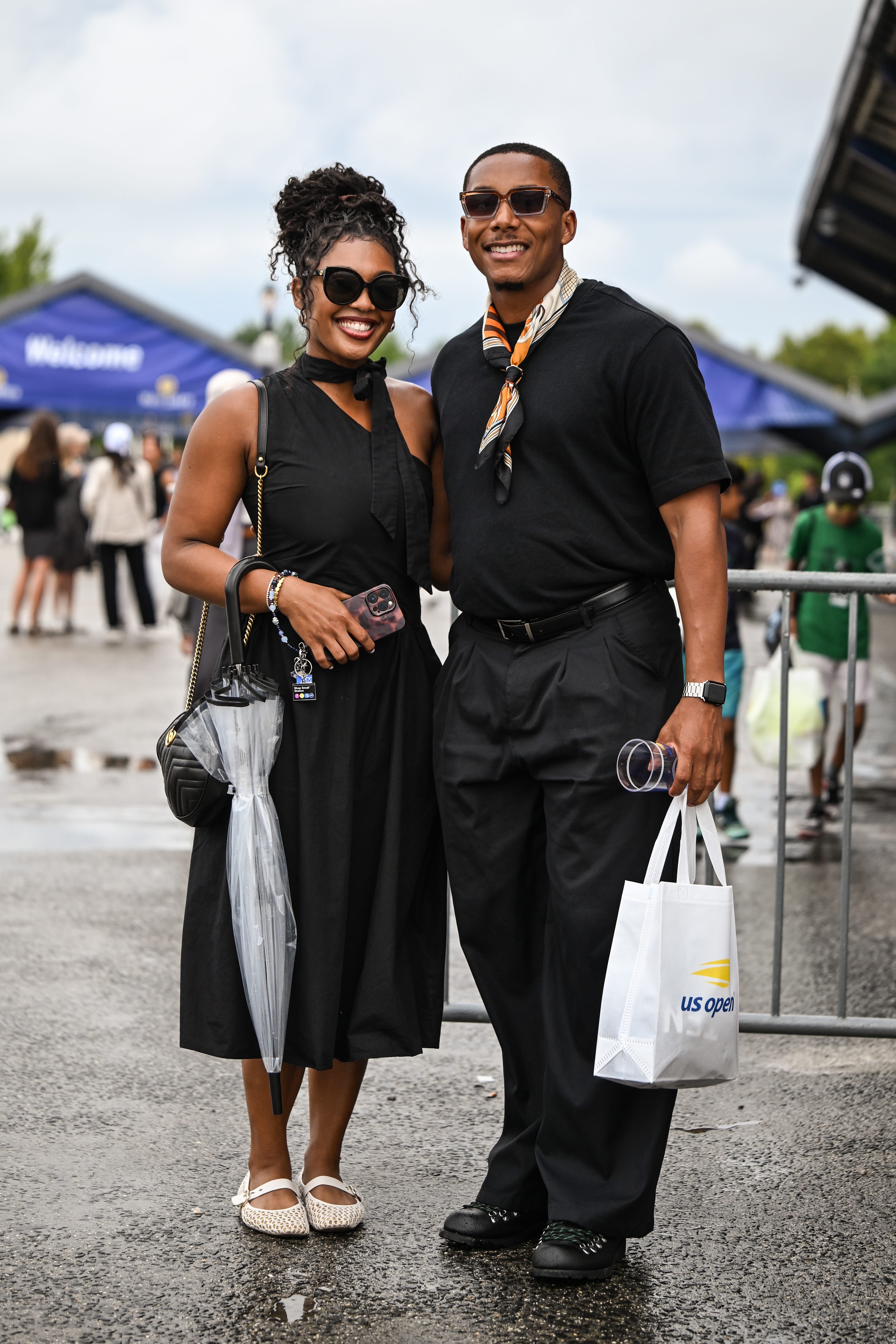 Two stylish spectators in black outfits pose outdoors at a US Open event with a branded shopping bag.
