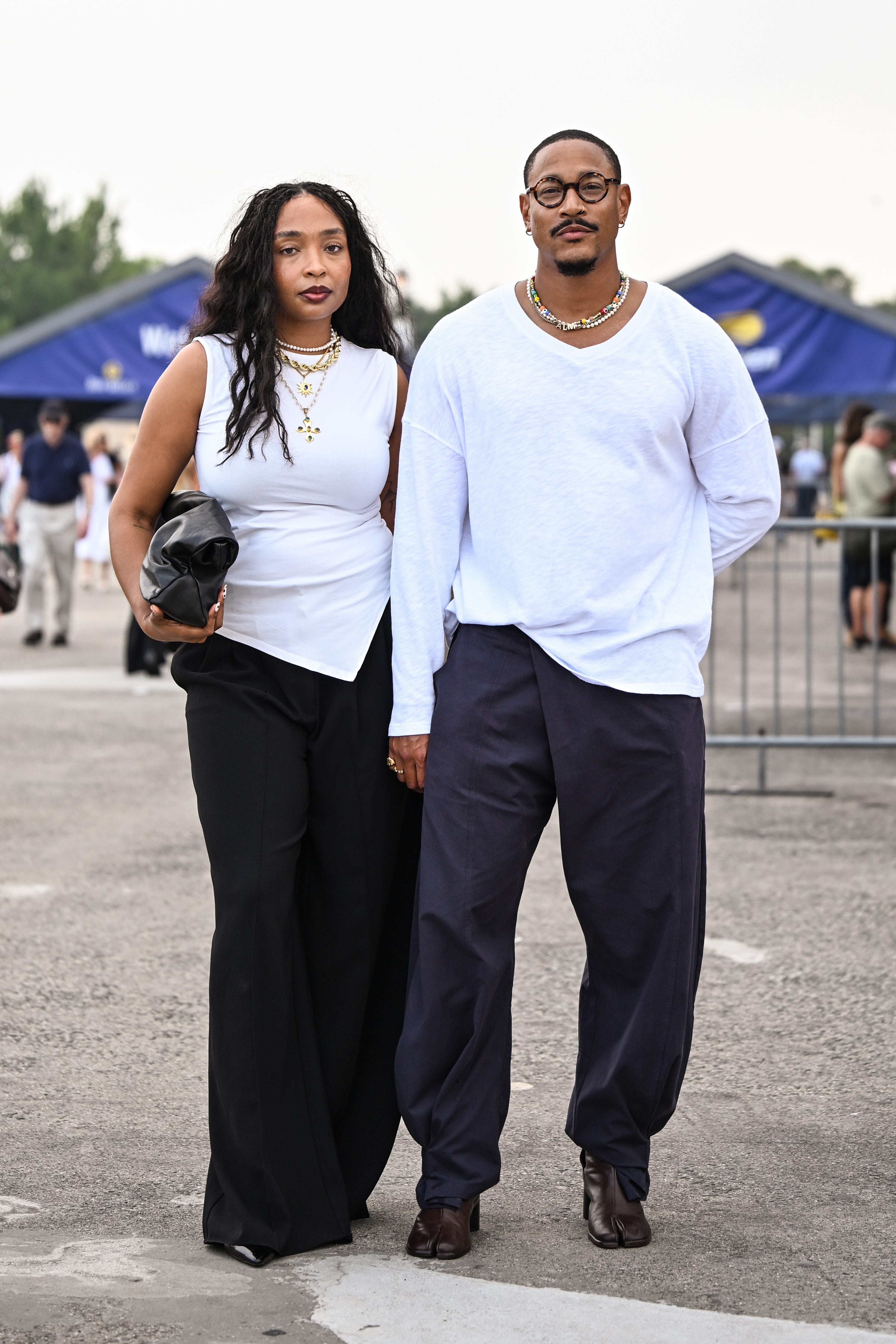 Two people in white tops and dark trousers stand holding hands at an outdoor event entrance.