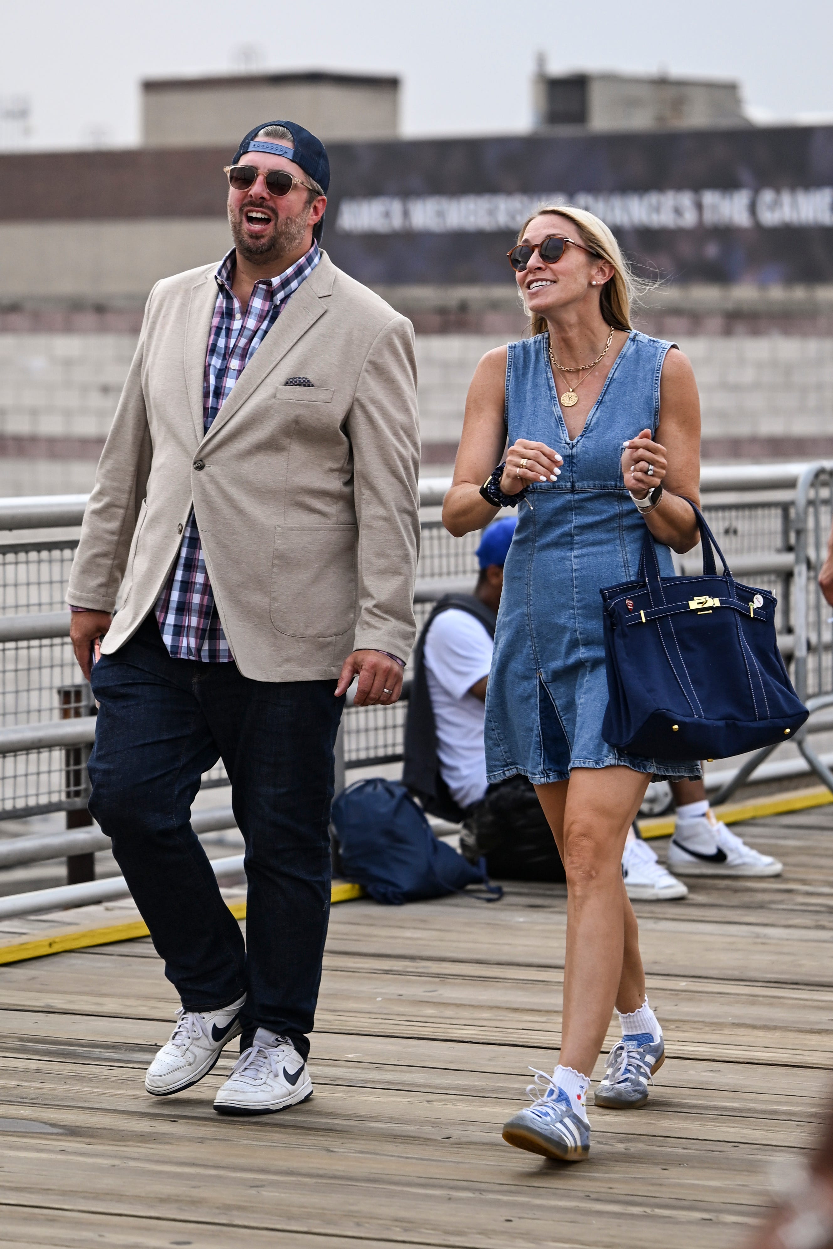 Two people walk on a wooden boardwalk wearing sunglasses and casual dressy outfits.