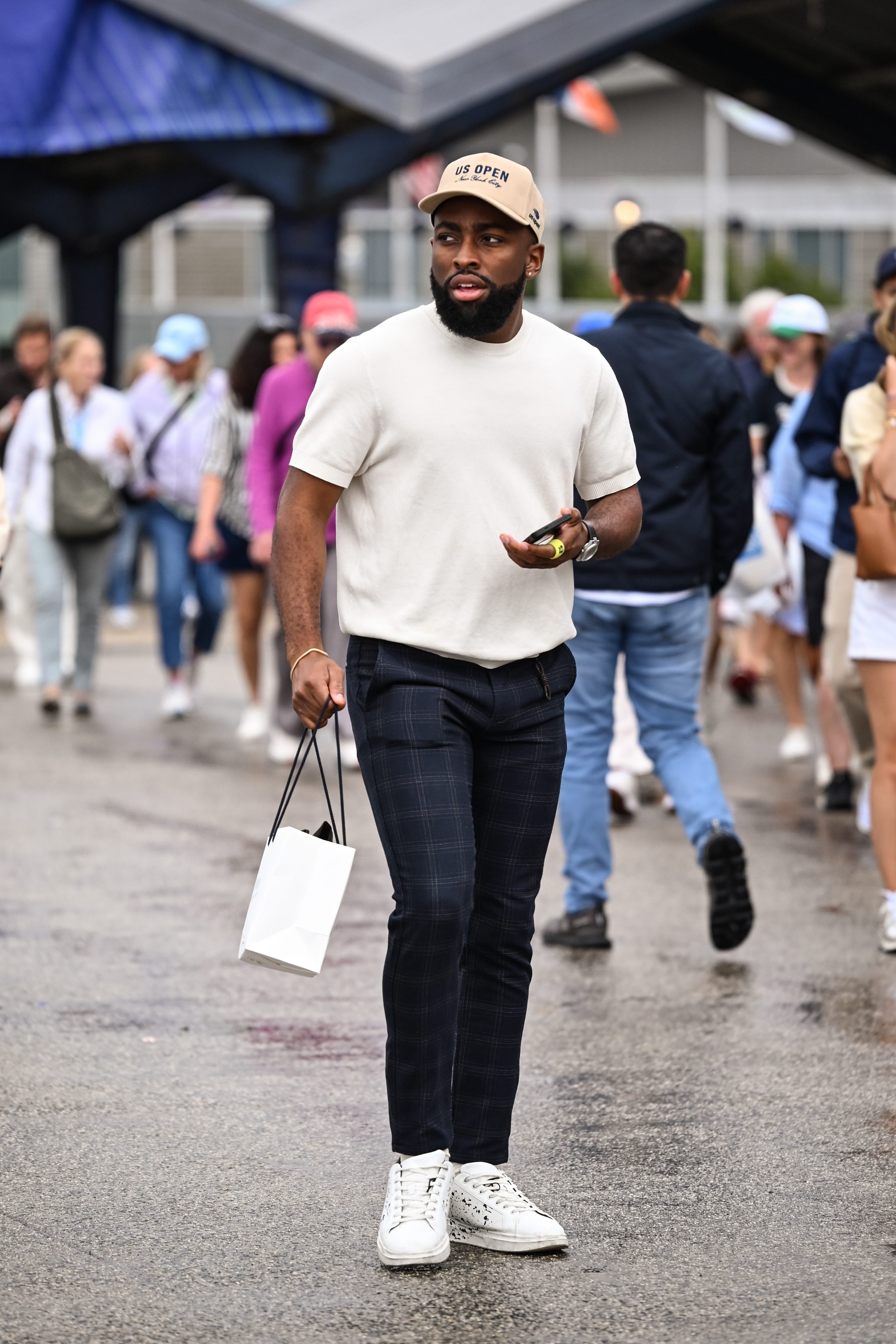 Person wearing a US OPEN cap walks through a rainy crowd carrying a small white shopping bag.