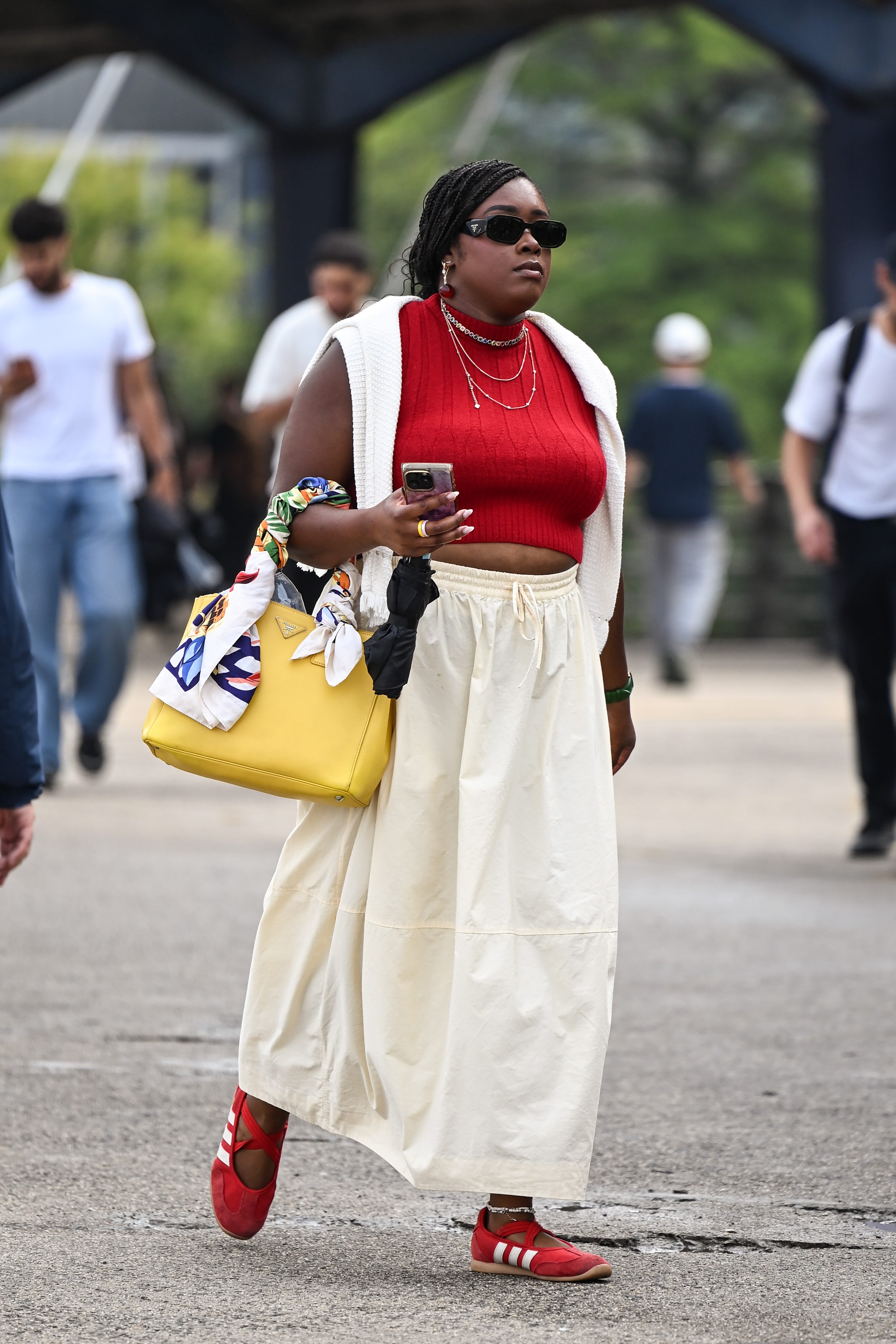 Person in sunglasses walks outdoors wearing a red top, cream skirt, red shoes, and carrying a yellow handbag.