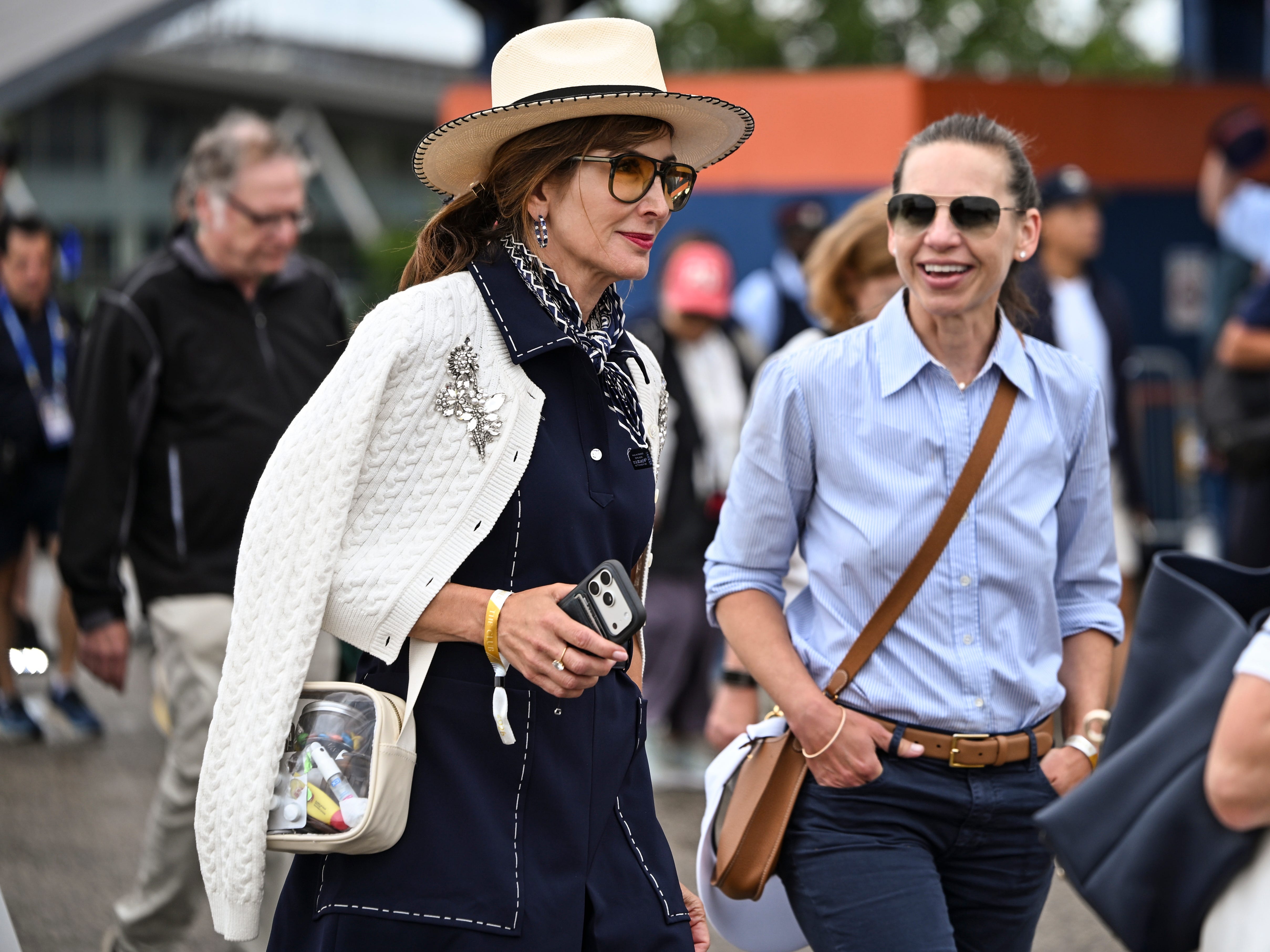 Two stylish attendees wearing sunglasses walk through a crowded outdoor event area, one holding a smartphone.
