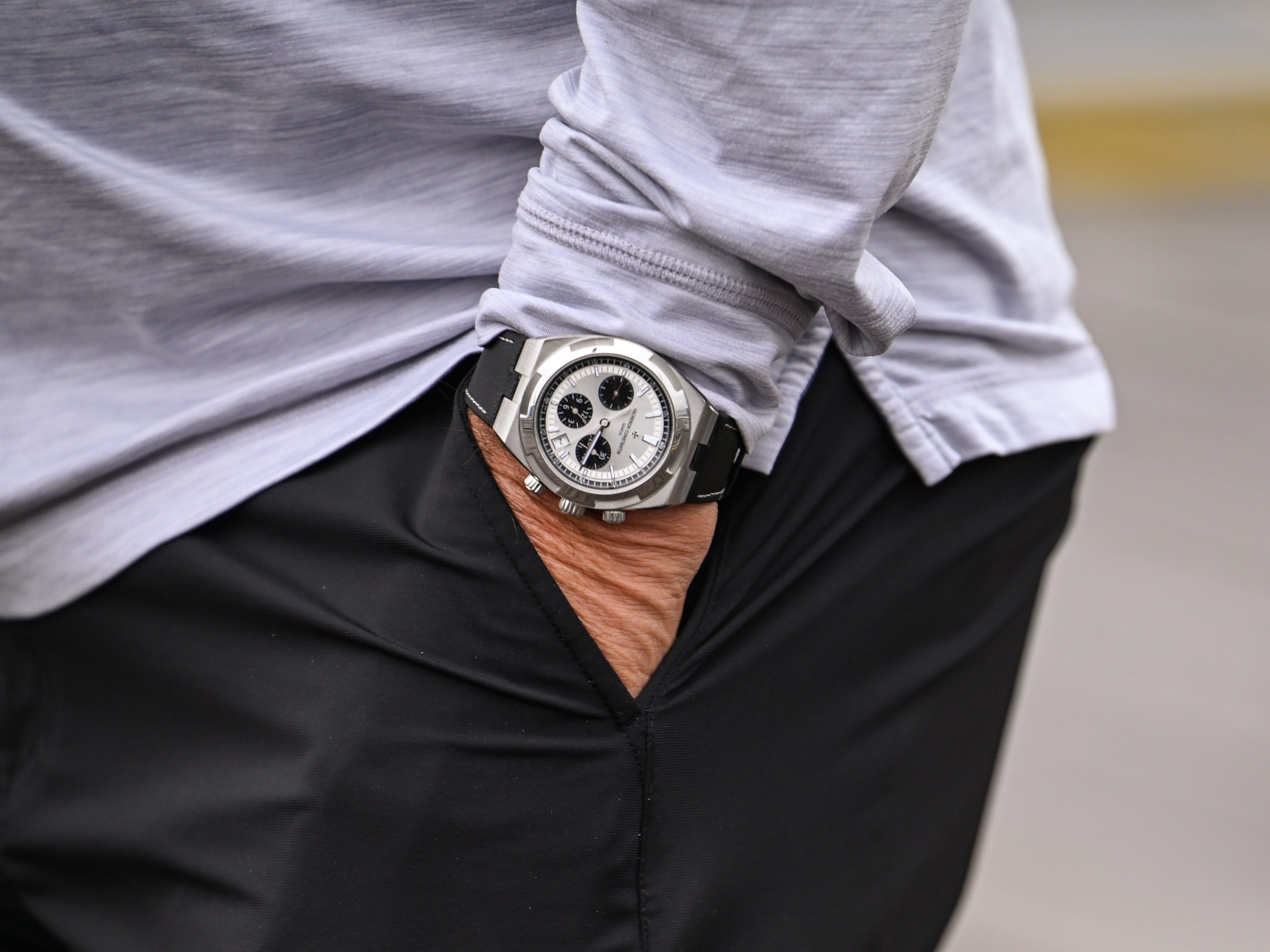Close-up of a chronograph wristwatch on a wrist beside black pants and a light gray long-sleeve shirt.