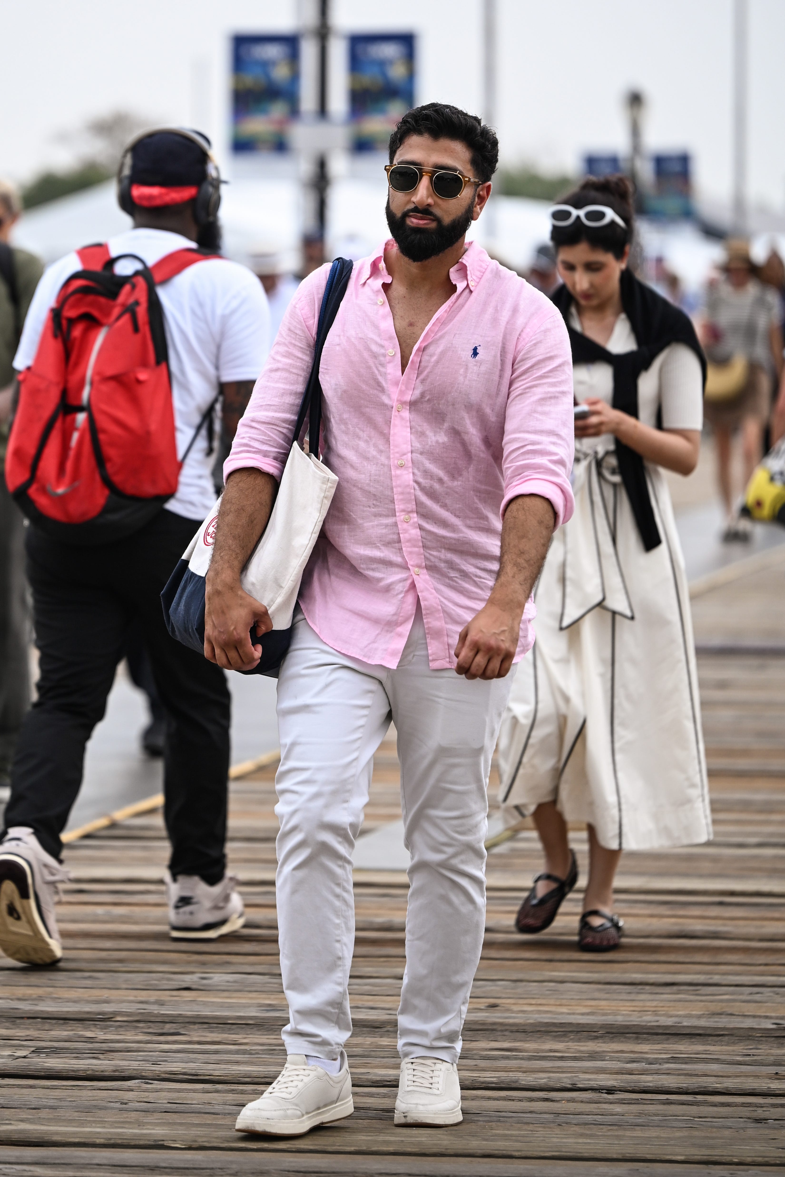 Person in a pink button-down shirt and white pants walks on a wooden boardwalk among pedestrians.