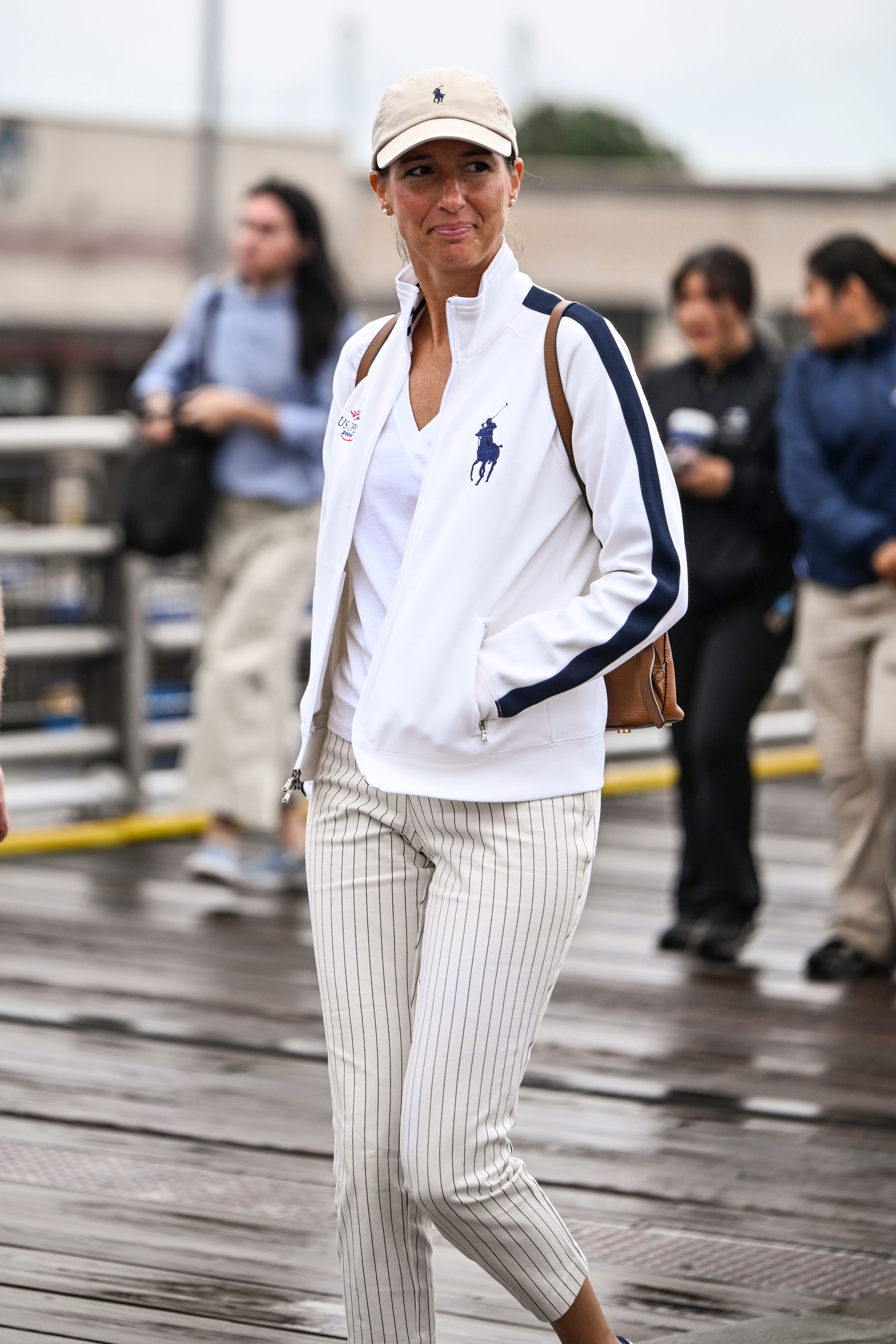 Person in white Polo Ralph Lauren jacket and striped trousers walks on a wet boardwalk in an urban setting.