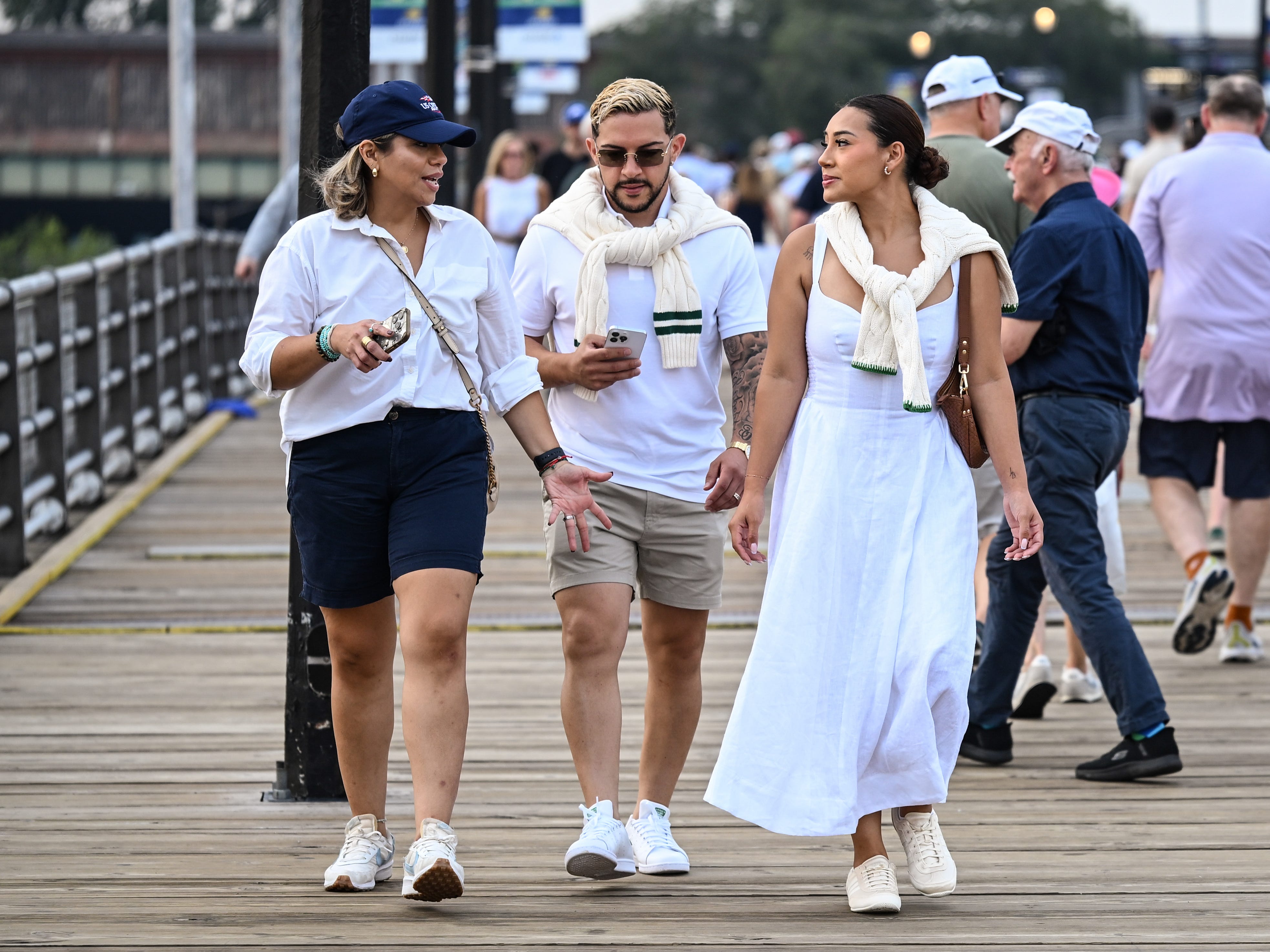 Three casually dressed people walk together on a crowded wooden pier near railings and streetlights.